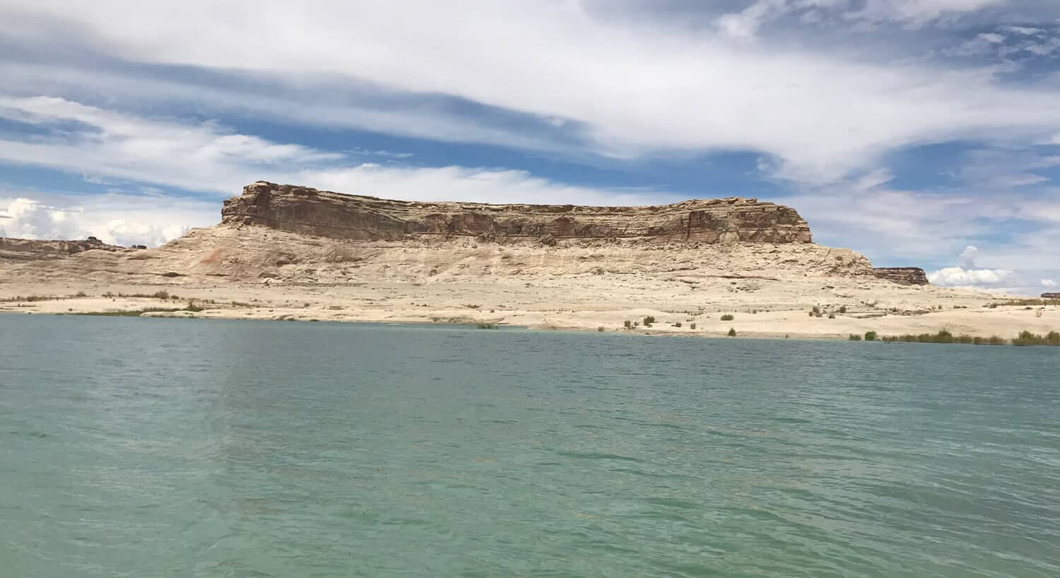 View across clear water toward tan rock formation under bright blues sky with fluffy white clouds.