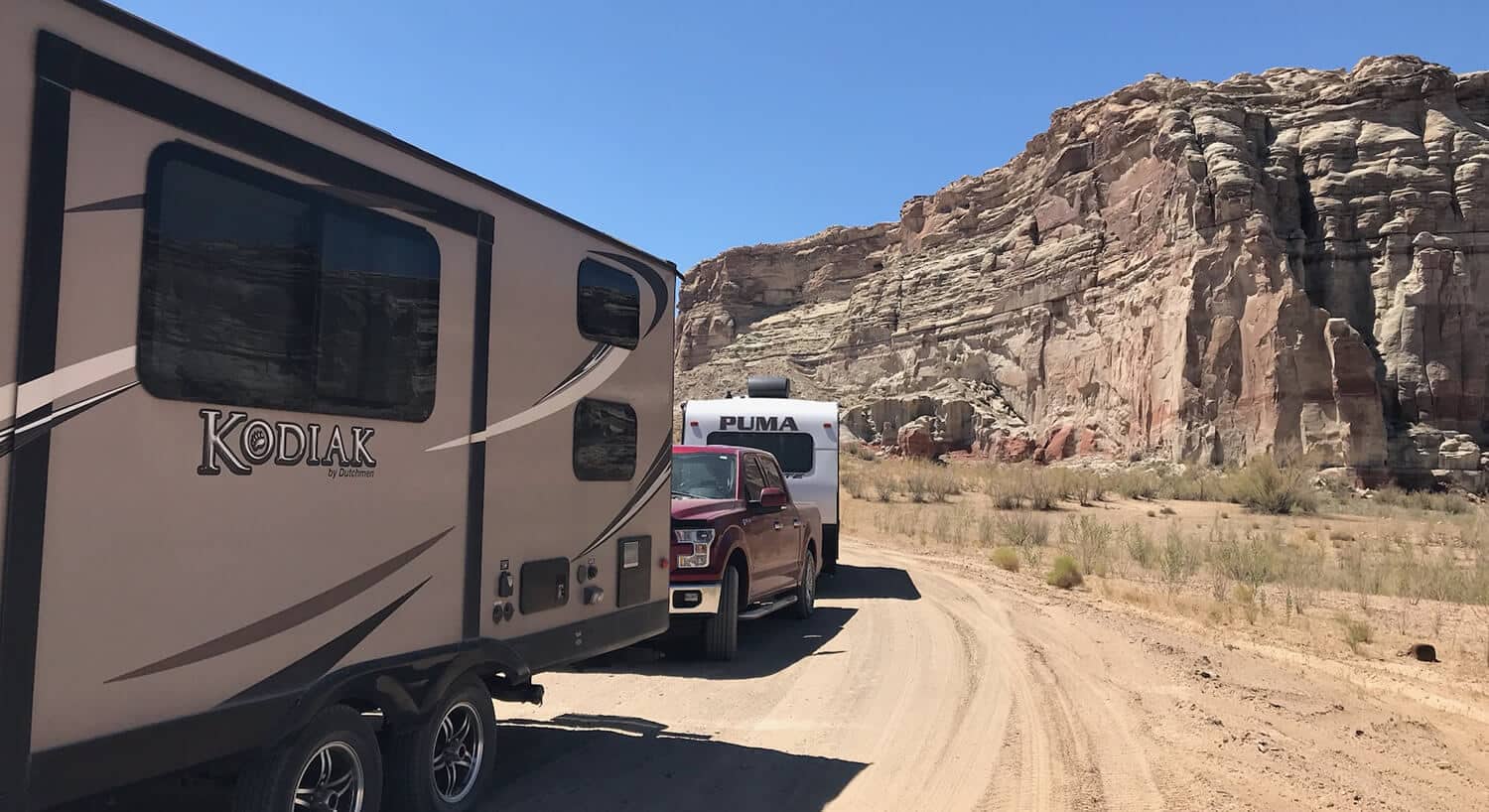 Tan Kodiak camper on dirt road with red truck pulling white Puma caper with tan rocks on the side