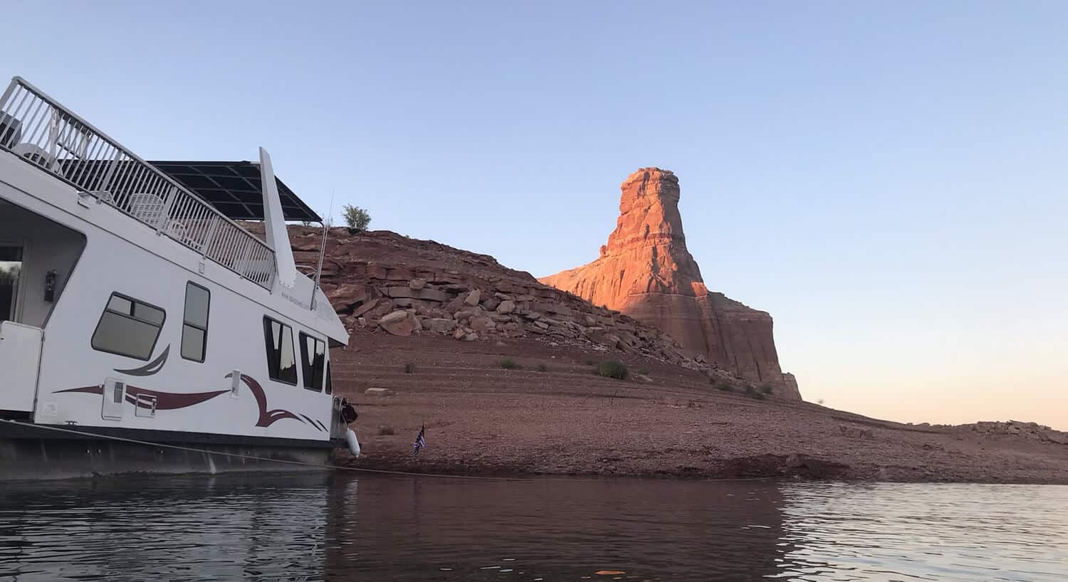White houseboat docked on beach with balcony with white lawn chairs with tall cathedral rock formation in distance