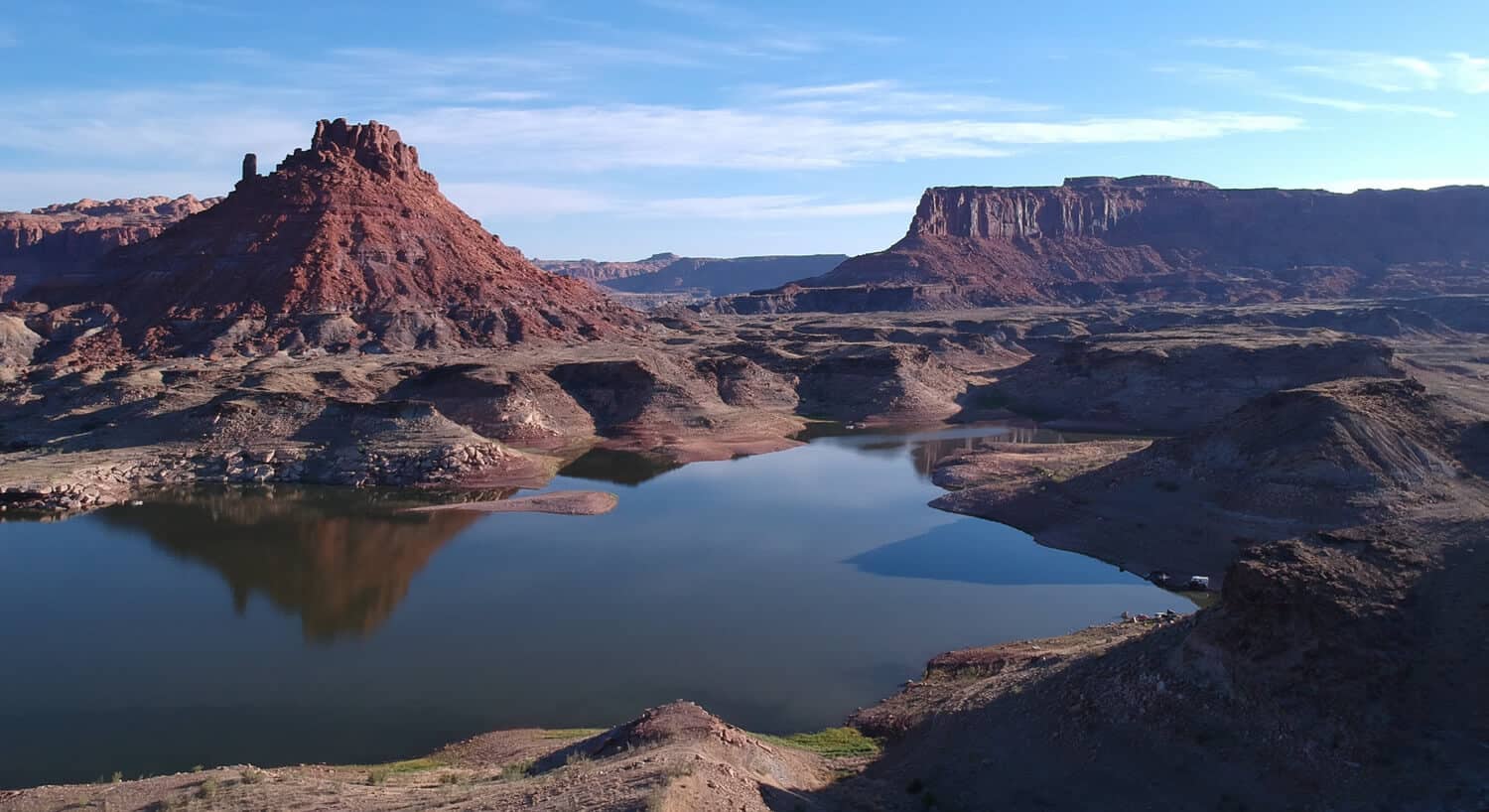 Red rock formations and mounds of dirt reflecting off water in front under blue sky with fluffy white clouds