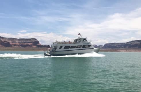 white tour boat with American flag speeding along water with rippling waves behind, red rock formations in the distance