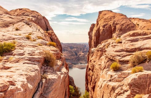 view of water through a narrow opening between red rock formations with schrubs growing, additional rock formations in distance, low handgin fluffy clouds