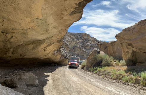 red Ford truck pulling tan camper along dirt in between rock formations and ledges